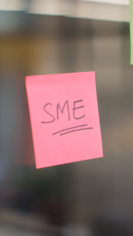 Vertical-Video-Close-Up-Of-Woman-Putting-Sticky-Note-With-SME-Written-On-It-Onto-Transparent-Screen-In-Office